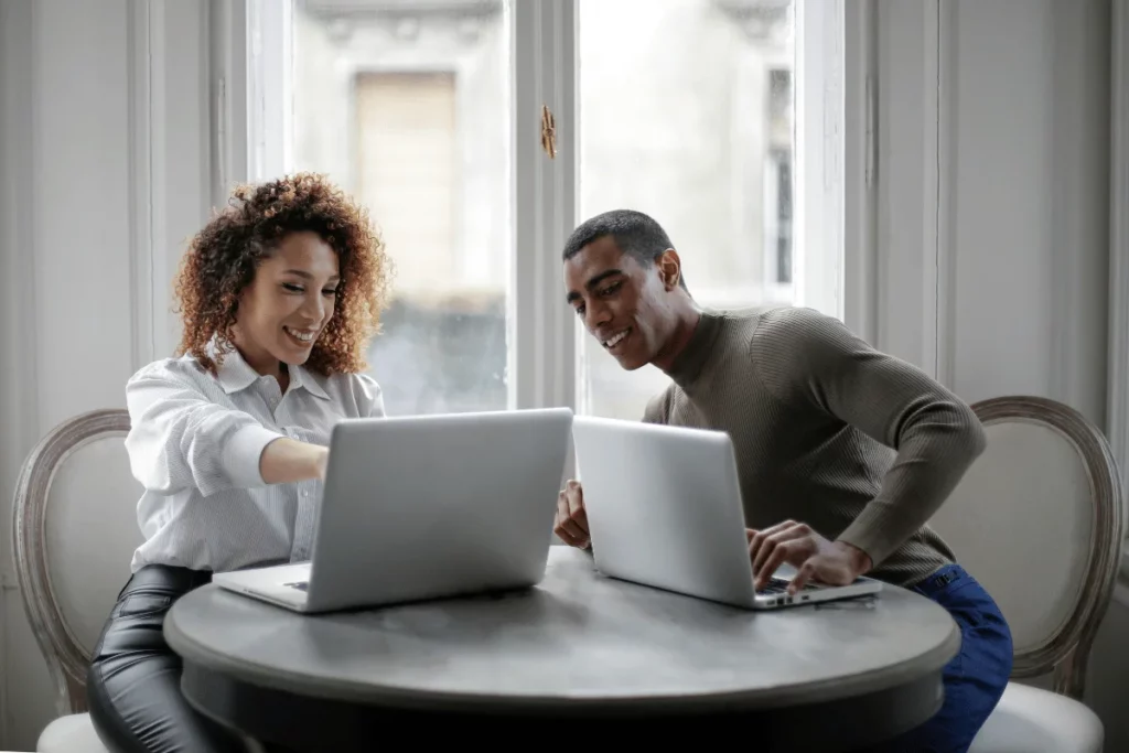 Two colleagues working side by side with laptops, smiling and discussing their work.