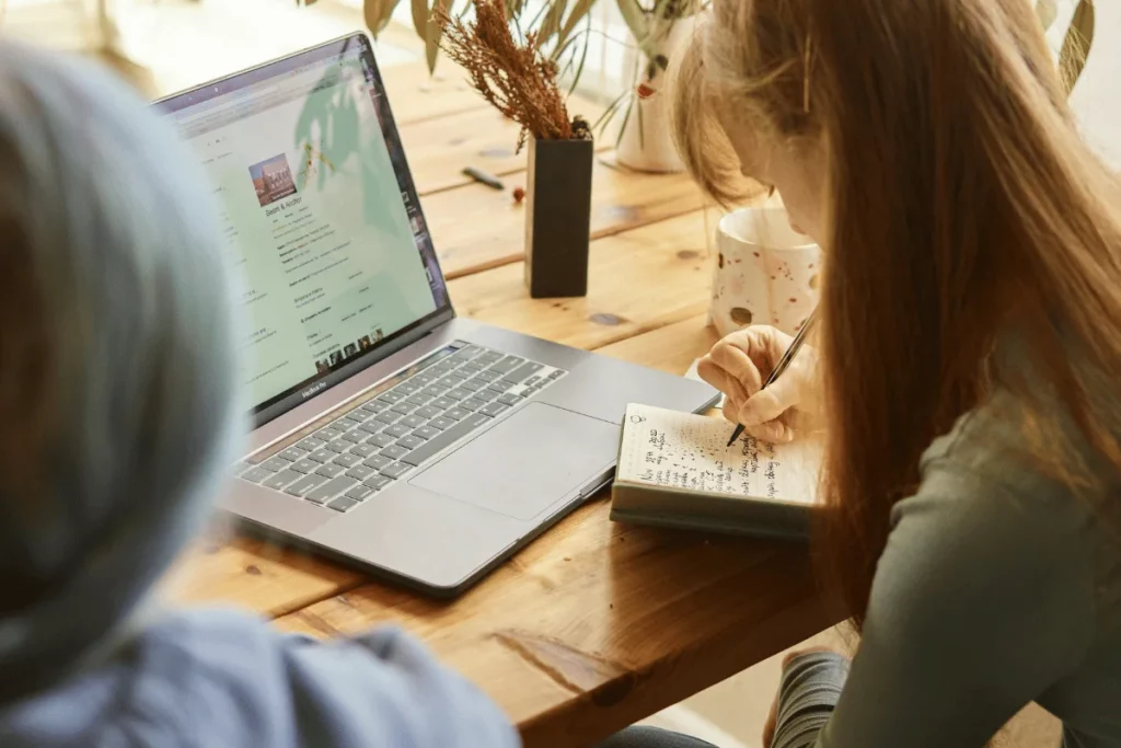 Woman taking handwritten notes while researching on a laptop.