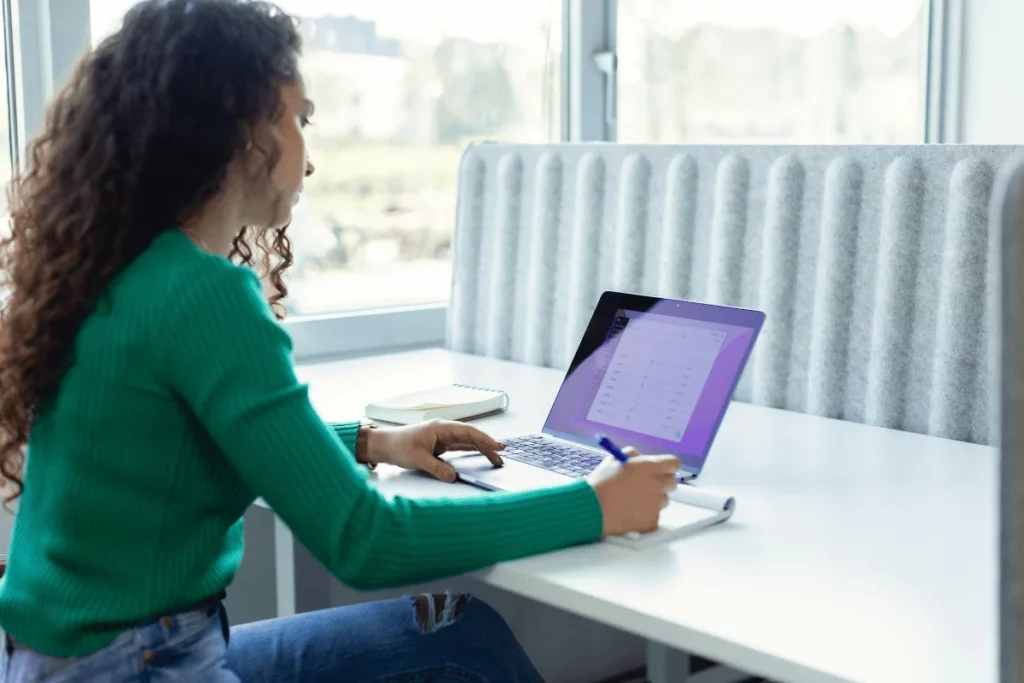 Woman in a green sweater working on a laptop in a bright, modern workspace.