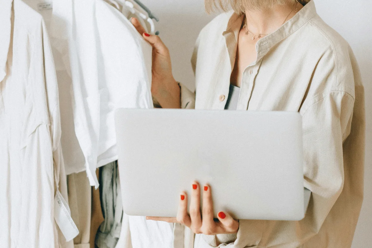 Woman in beige shirt holding a laptop and browsing through white clothes on hangers.