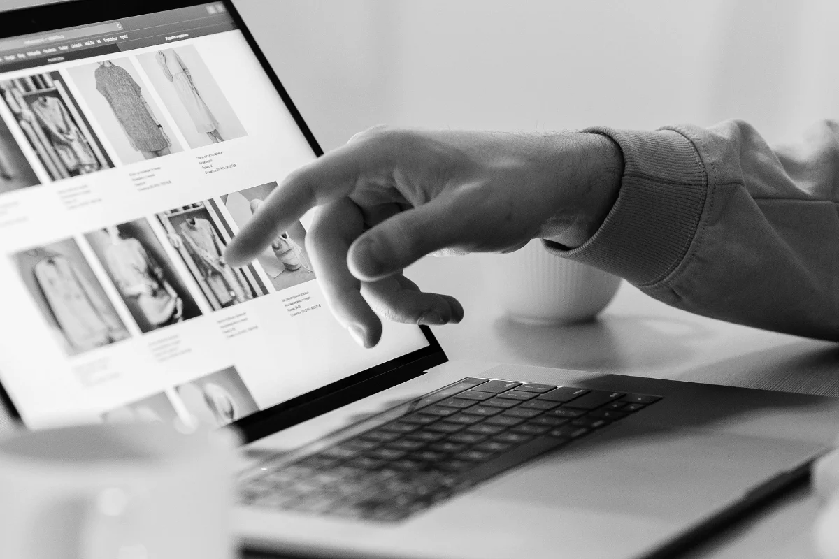 Hand pointing at fashion clothing items displayed on a laptop screen.