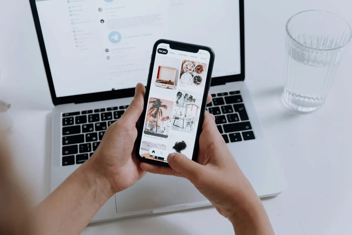 Person scrolling through interior design images on a smartphone, with a laptop in the background.
