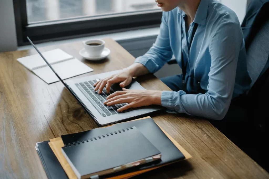 Professional typing on a laptop at a desk with notebooks and coffee nearby.