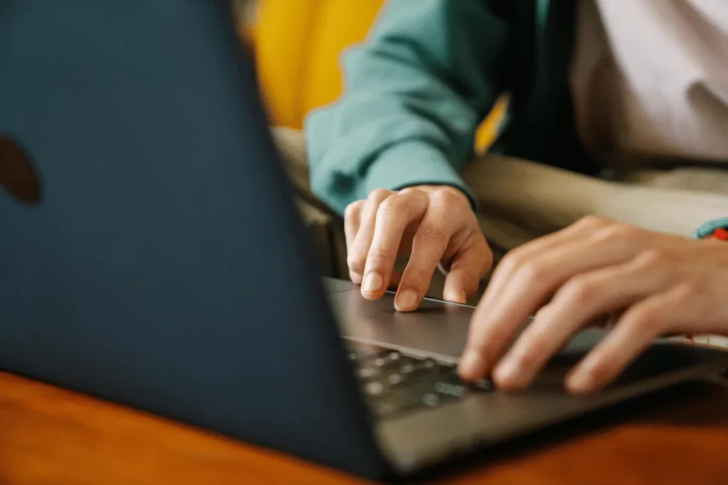 A person types on a laptop keyboard, resting on a wooden table, with a green sweater and casual beige pants.