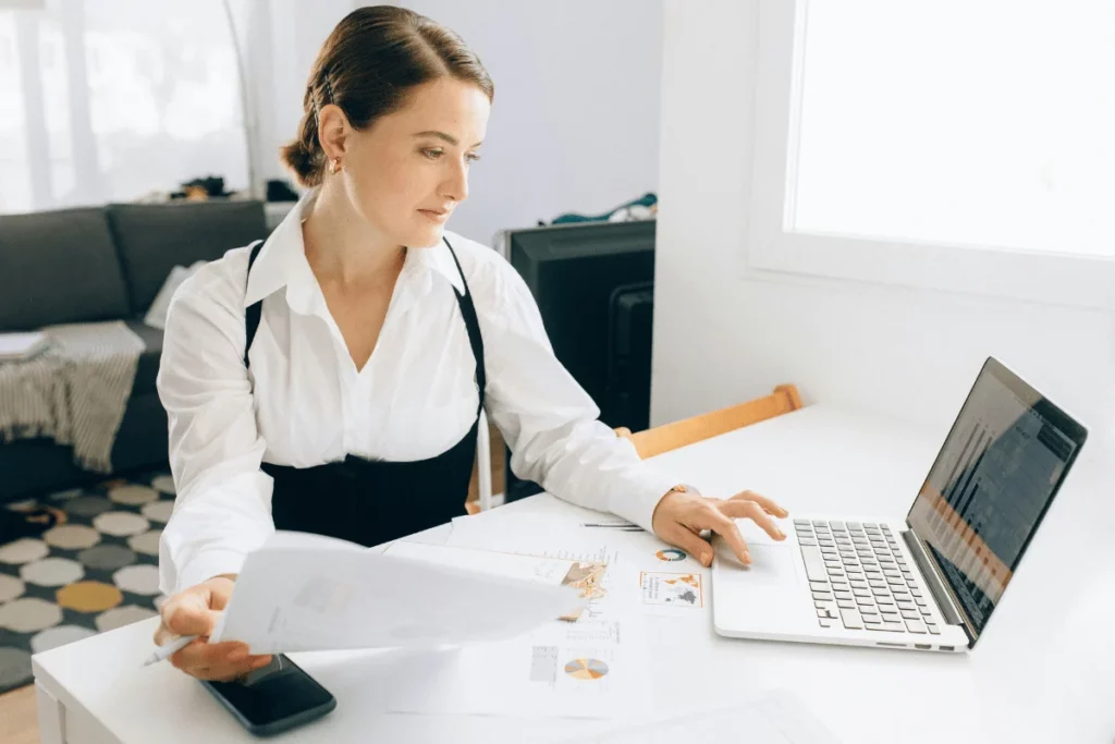 A woman in a white shirt and black apron reviews documents and works on a laptop at a bright, modern office desk.
