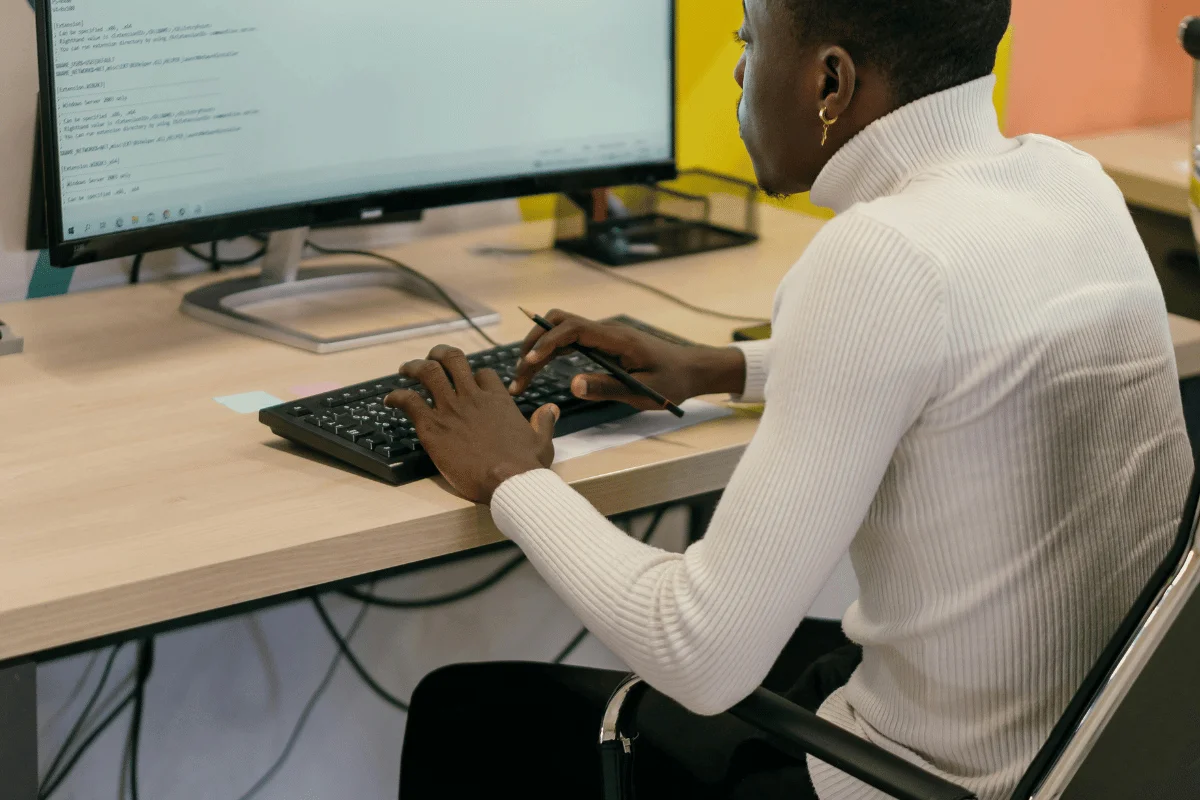 A person in a white turtleneck types on a keyboard, focused on a large monitor displaying code. The setting conveys a work or study environment.