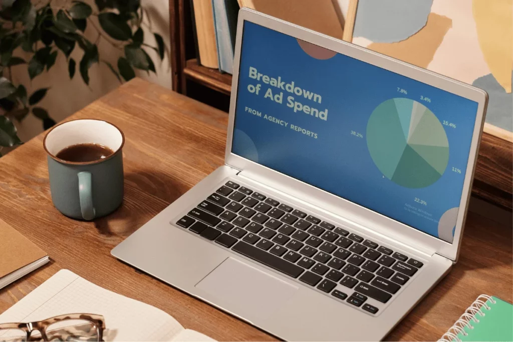 A laptop displaying a presentation on ad spend breakdown, next to a cup of coffee on a wooden desk.