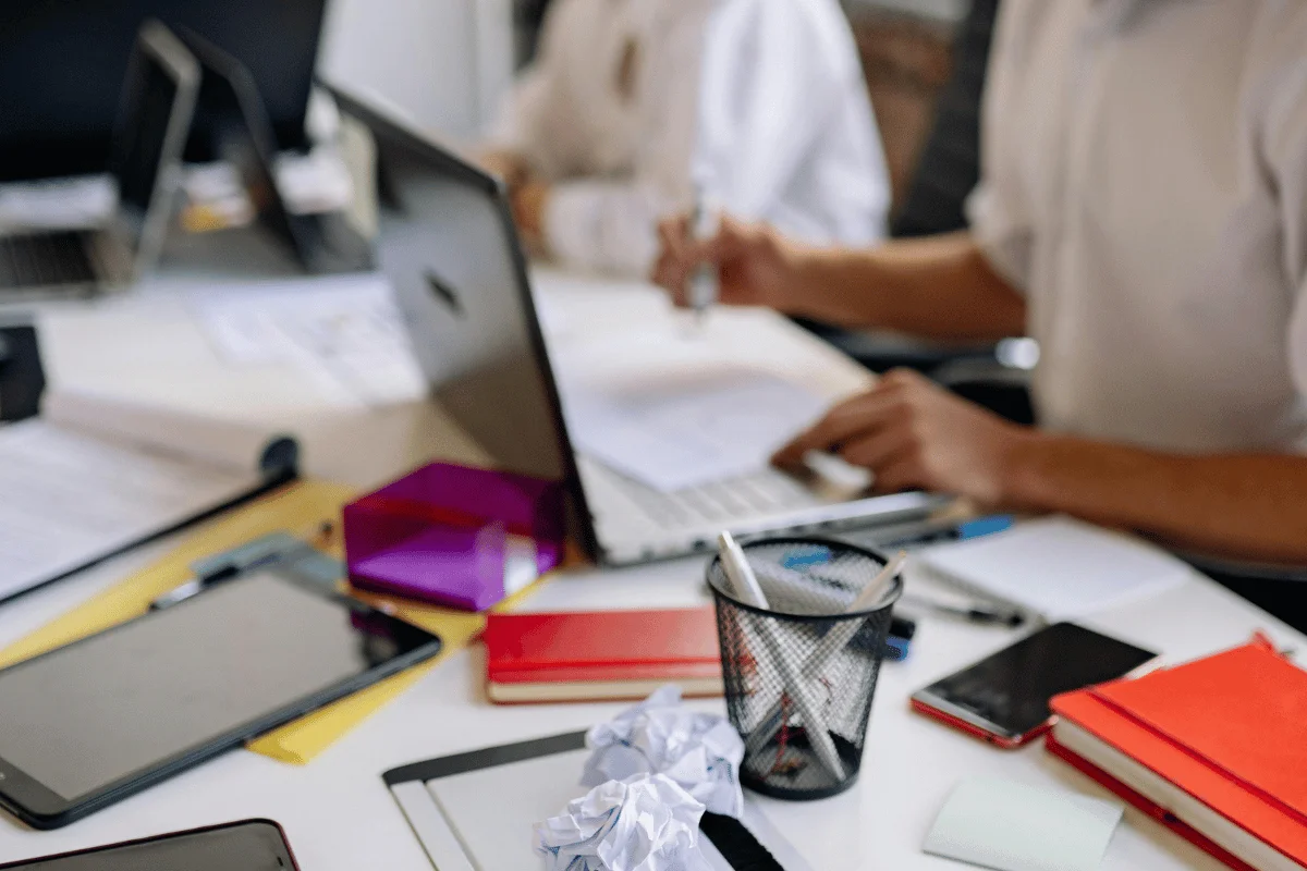 A workspace cluttered with laptops, notebooks, a trash can, and a hand writing on a document, indicating a busy brainstorming session.