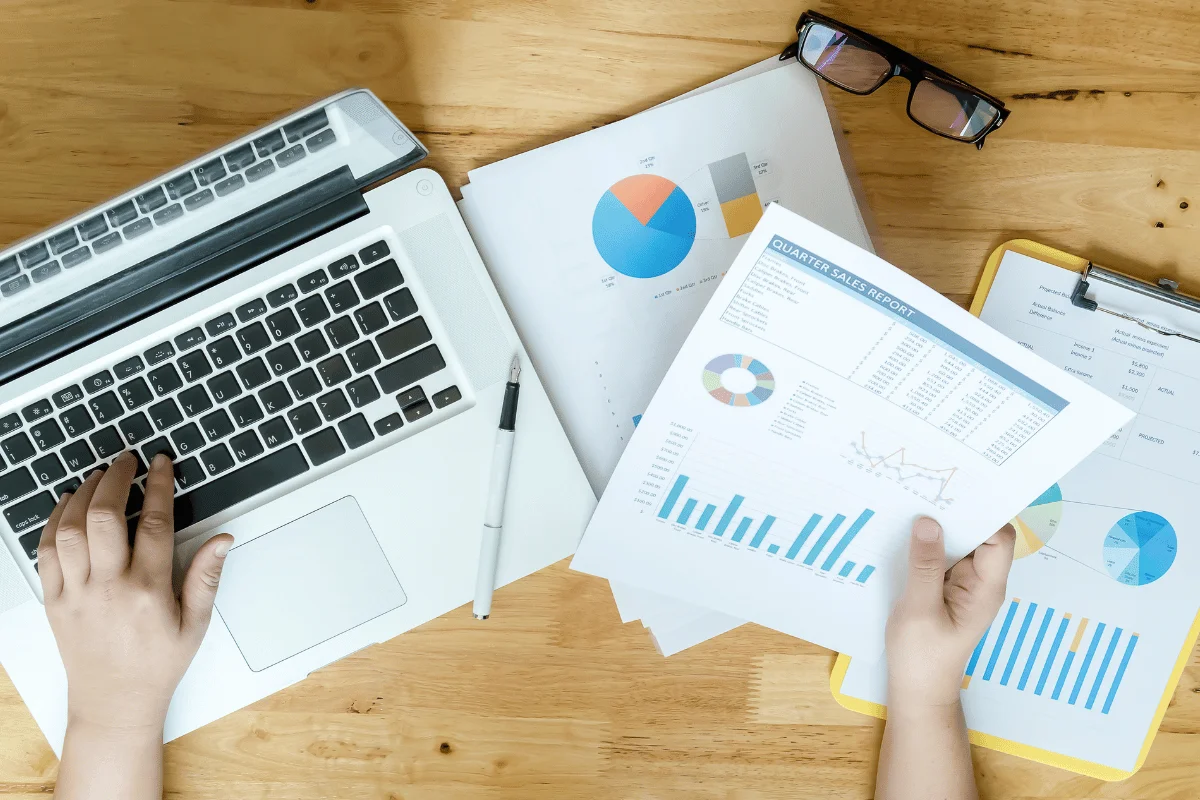 Person analyzing quarterly sales report charts next to an open laptop on desk.