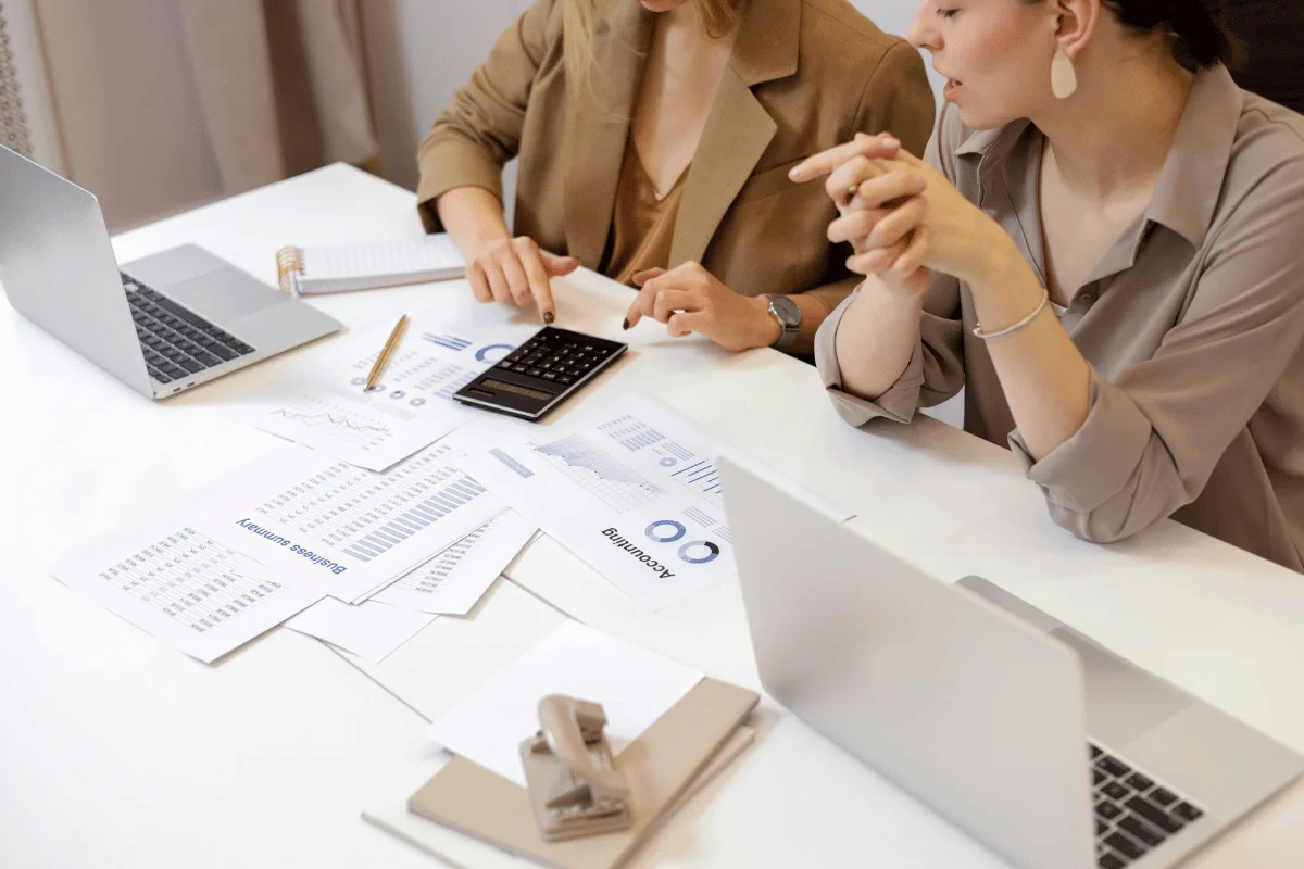 Two women collaborating on financial analysis with a calculator, charts, and laptops on the table.