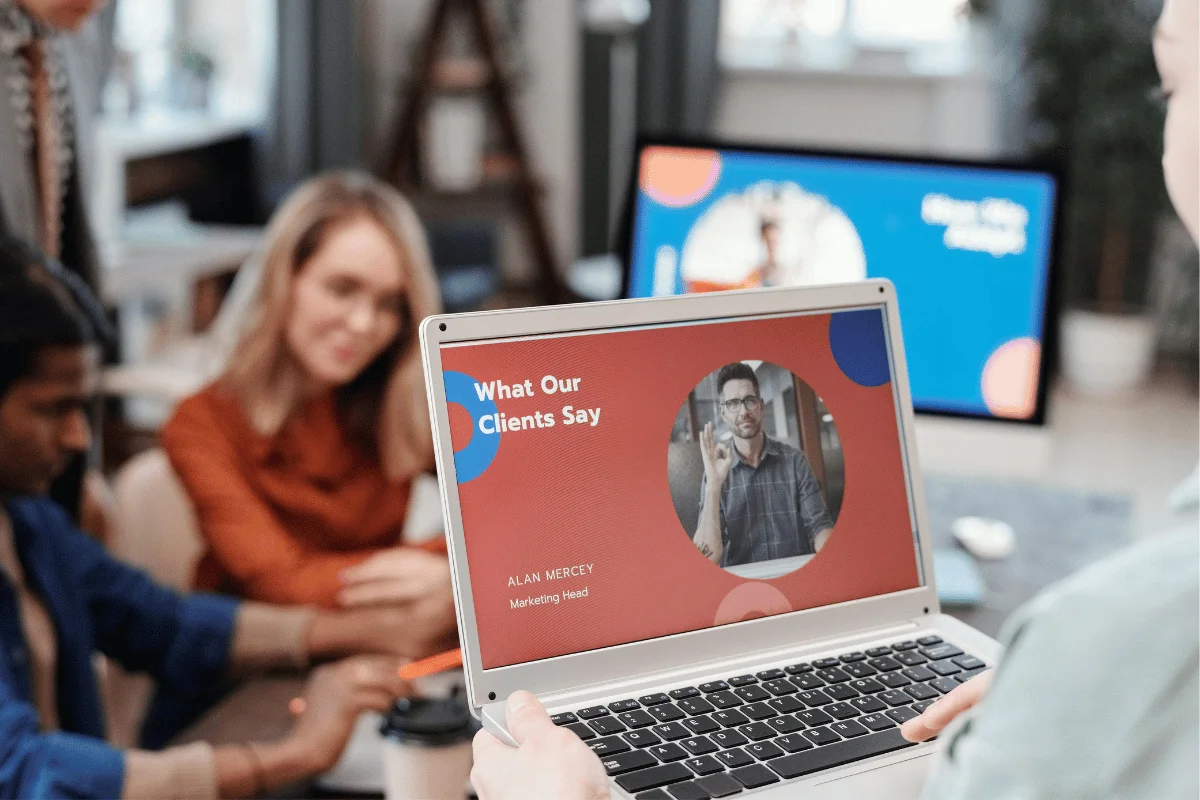 Close-up of a laptop displaying a testimonial slide, held by a person in a meeting room with colleagues discussing in the background.