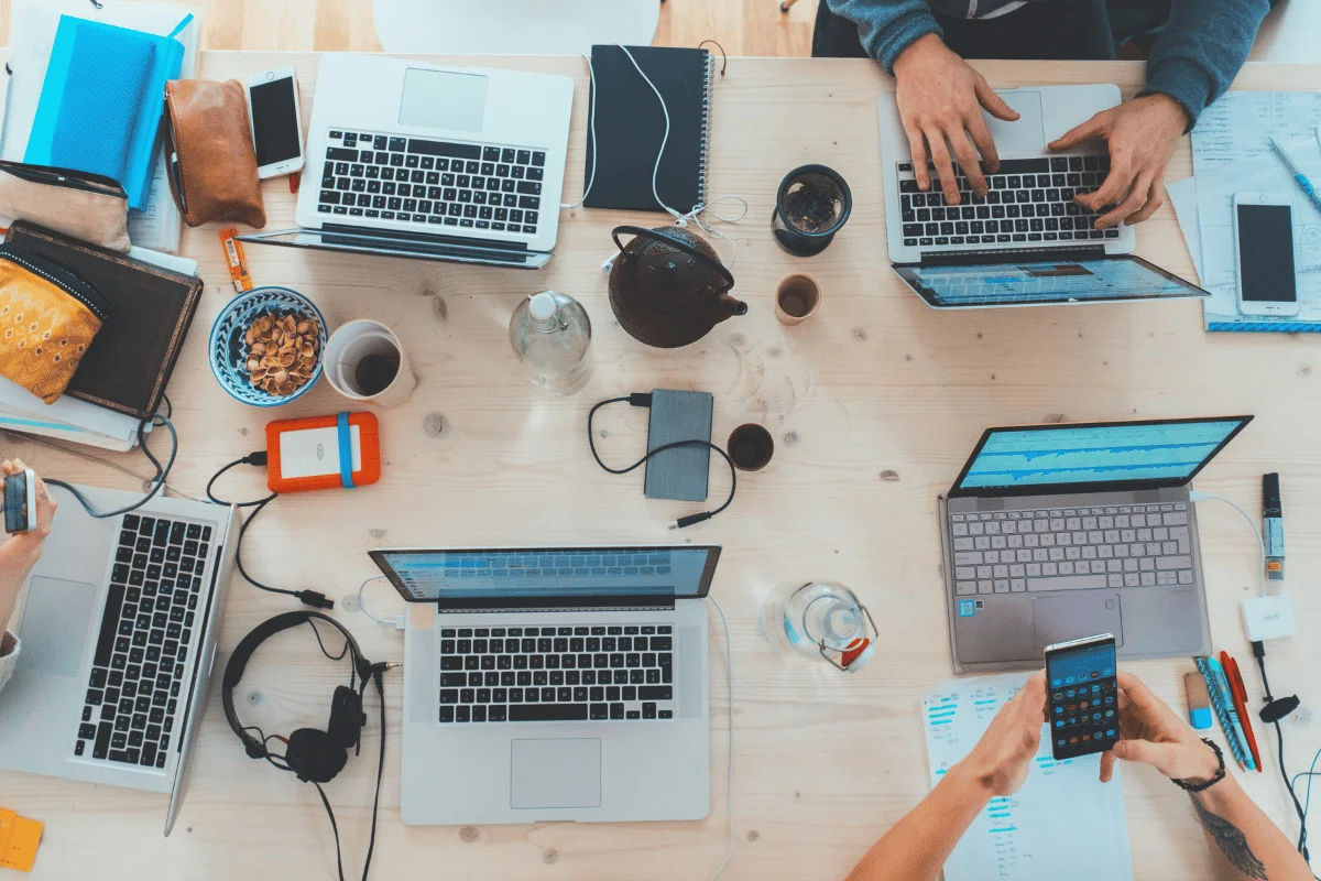 A group of people working together at a desk with multiple laptops, phones, and coffee cups.