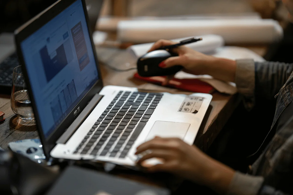 A person working on a laptop with a mouse, surrounded by papers and a glass of water.