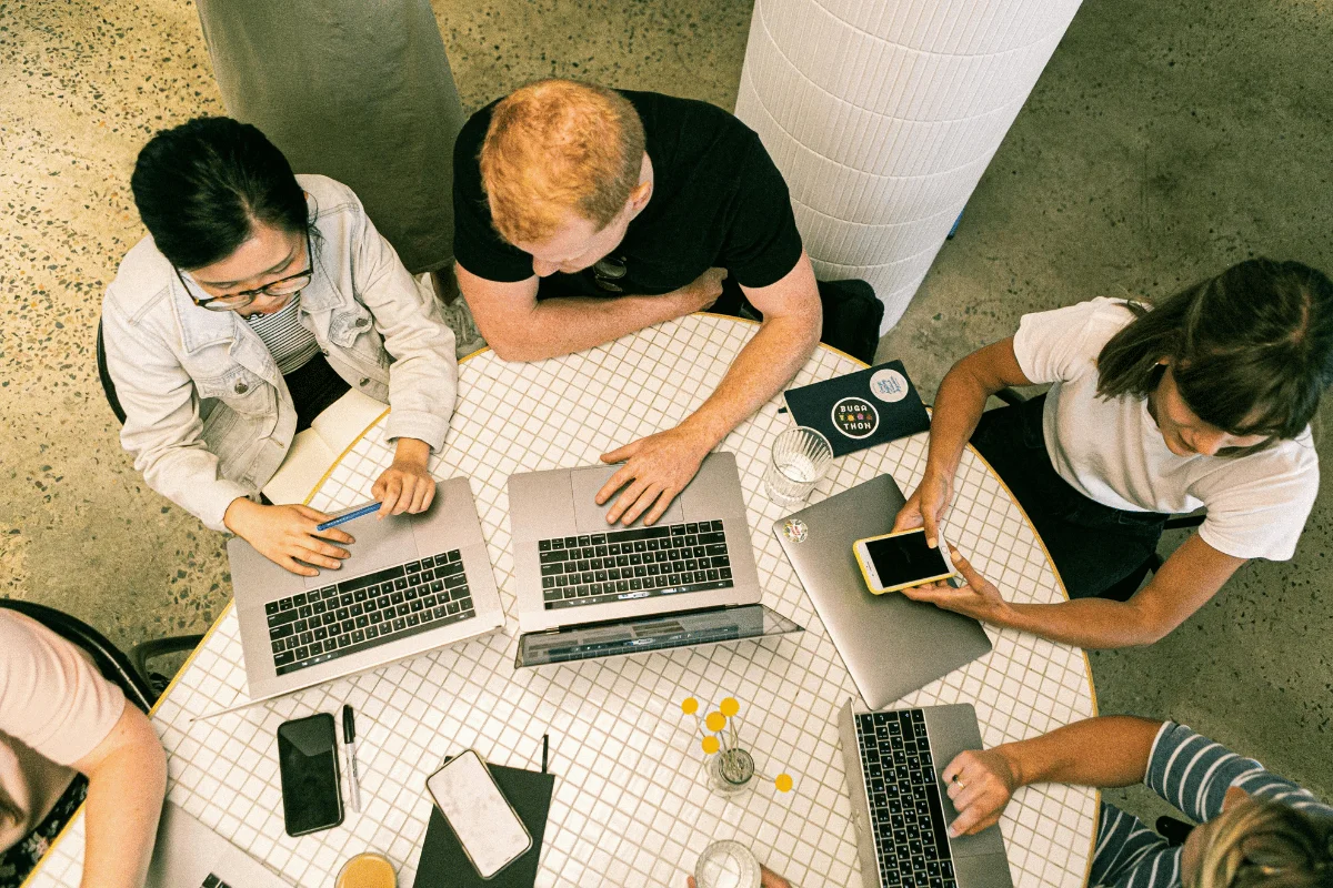 Overhead view of a group collaborating at a round table with laptops, notebooks, and a smartphone.