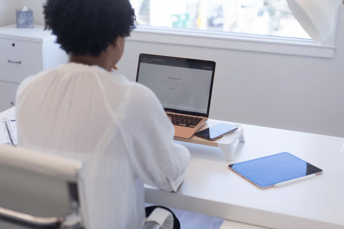 A woman with curly hair sits at a desk, focused on her laptop, surrounded by a tablet and smartphone, in a bright workspace.