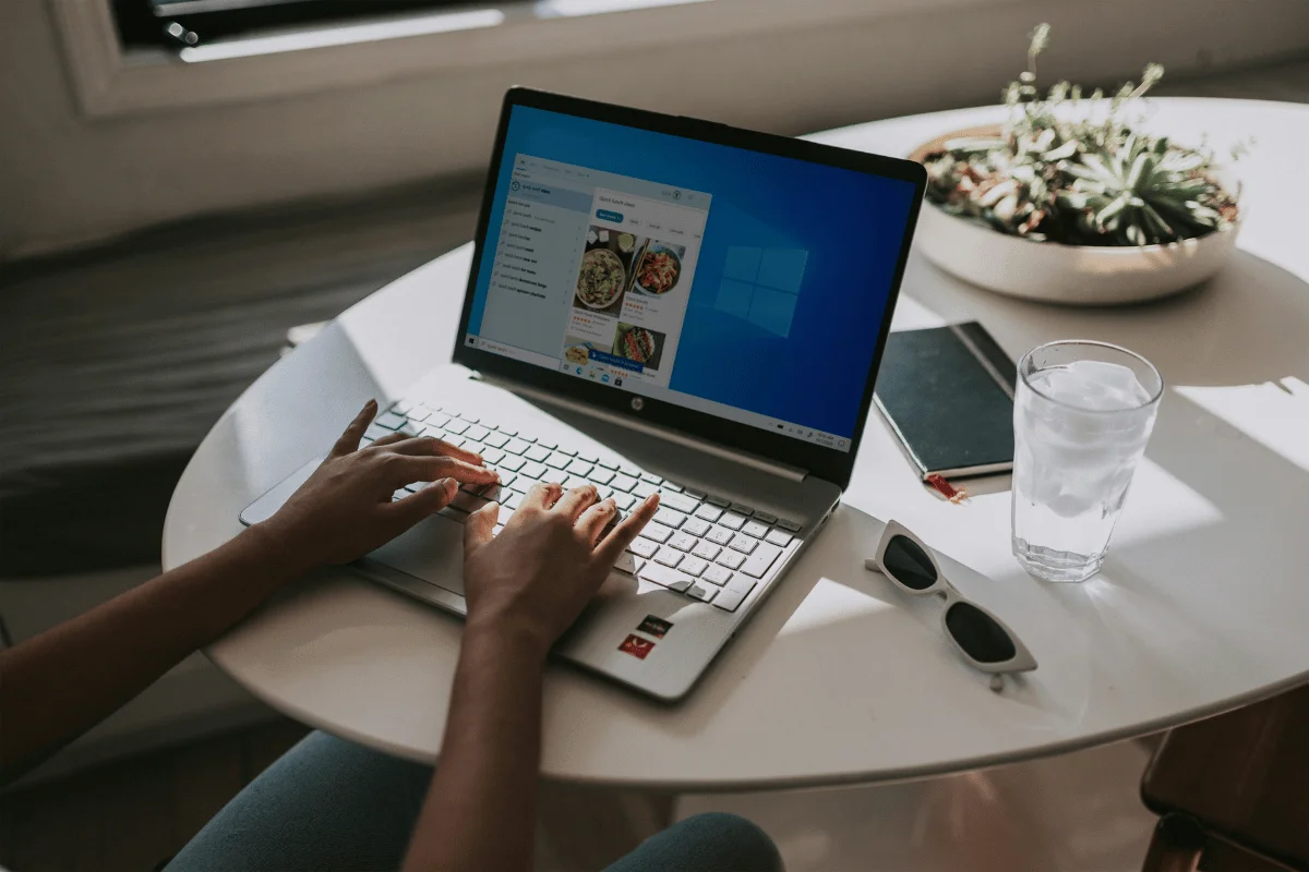 A person's hands typing on a laptop displaying a Windows interface, with a glass of water, a notebook, and sunglasses on the table.