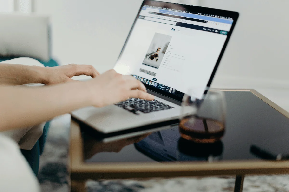 Individual typing on a laptop at a glass table, with a focus on their hands and a drink, in a contemporary home.
