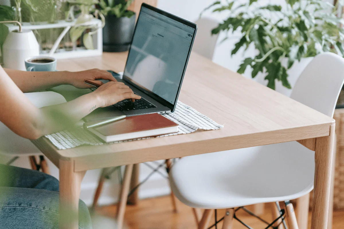 A person types on a laptop at a wooden table, with notebooks, a pen, and a coffee mug. 