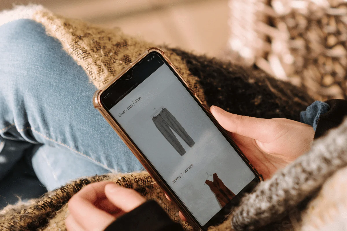 A person holds a smartphone, browsing clothing items including gray pants. They are wearing a knitted sweater and jeans, conveying a relaxed shopping mood.