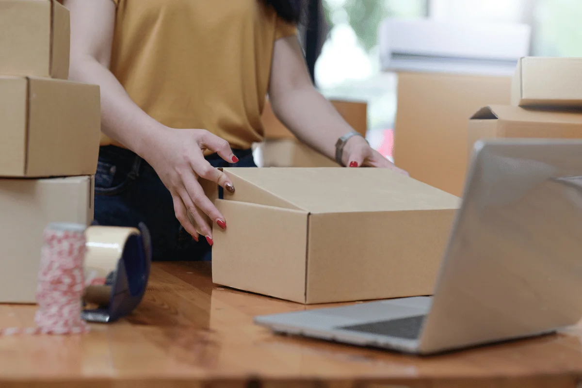 Woman handling a cardboard box on a packing table with a laptop nearby.