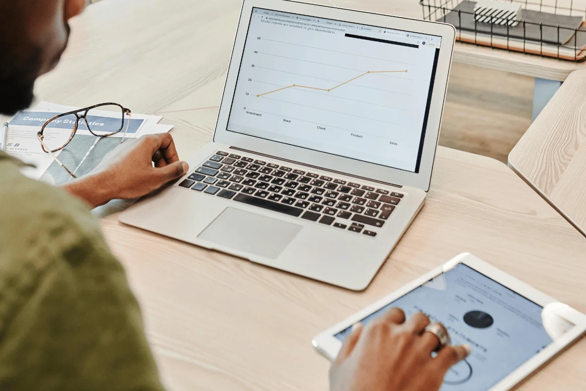 A person using a laptop showing a line graph and a tablet displaying a chart. Glasses and documents are on the desk, conveying a focused, analytical mood.