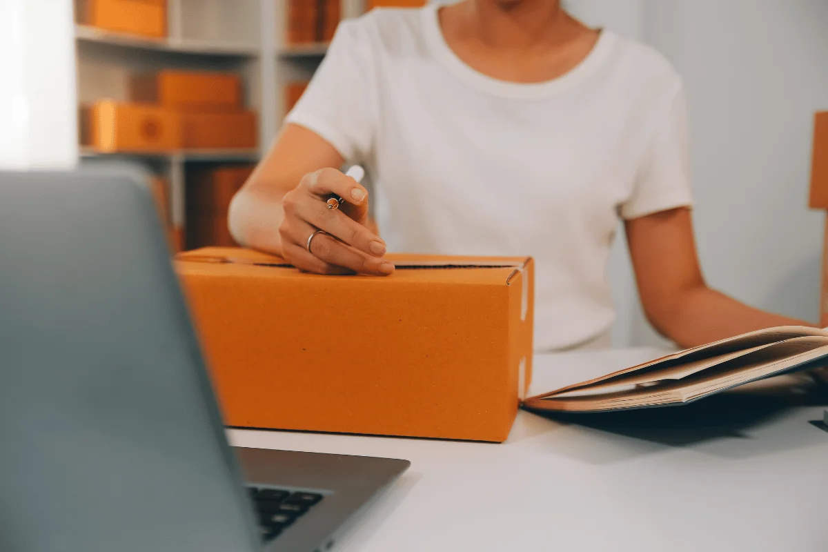A person in a white shirt uses a pen to write on an orange box while holding an open magazine beside a laptop.