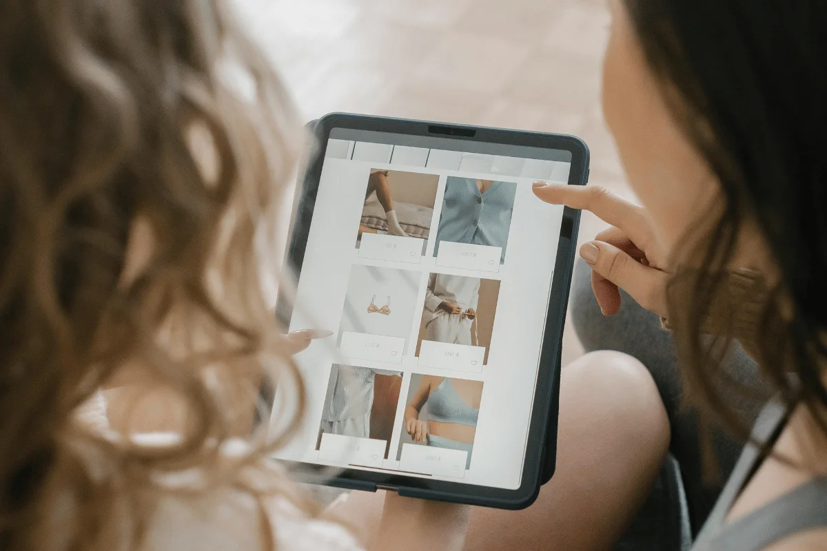 Two women are sitting together, reviewing clothing images on a tablet, pointing at pictures of bras and loungewear.