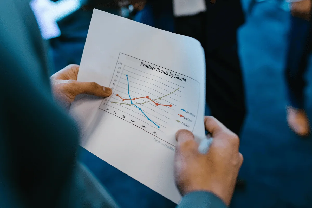 A close-up of a hand holding a printed graph titled "Product Trends by Month," showing data for desktops, laptops, and tablets.