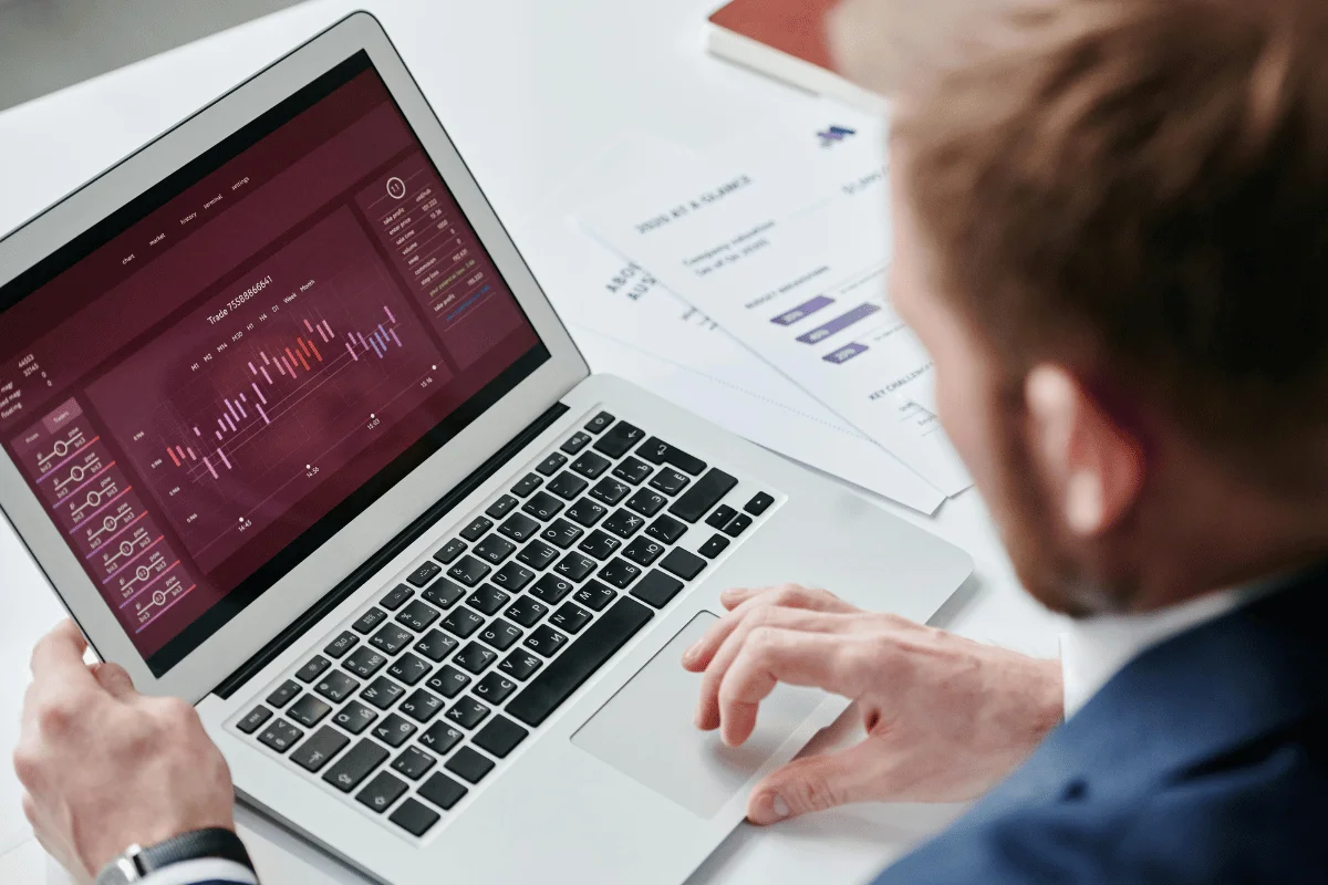 A person in a suit is using a laptop displaying financial charts and data, with papers in the background.