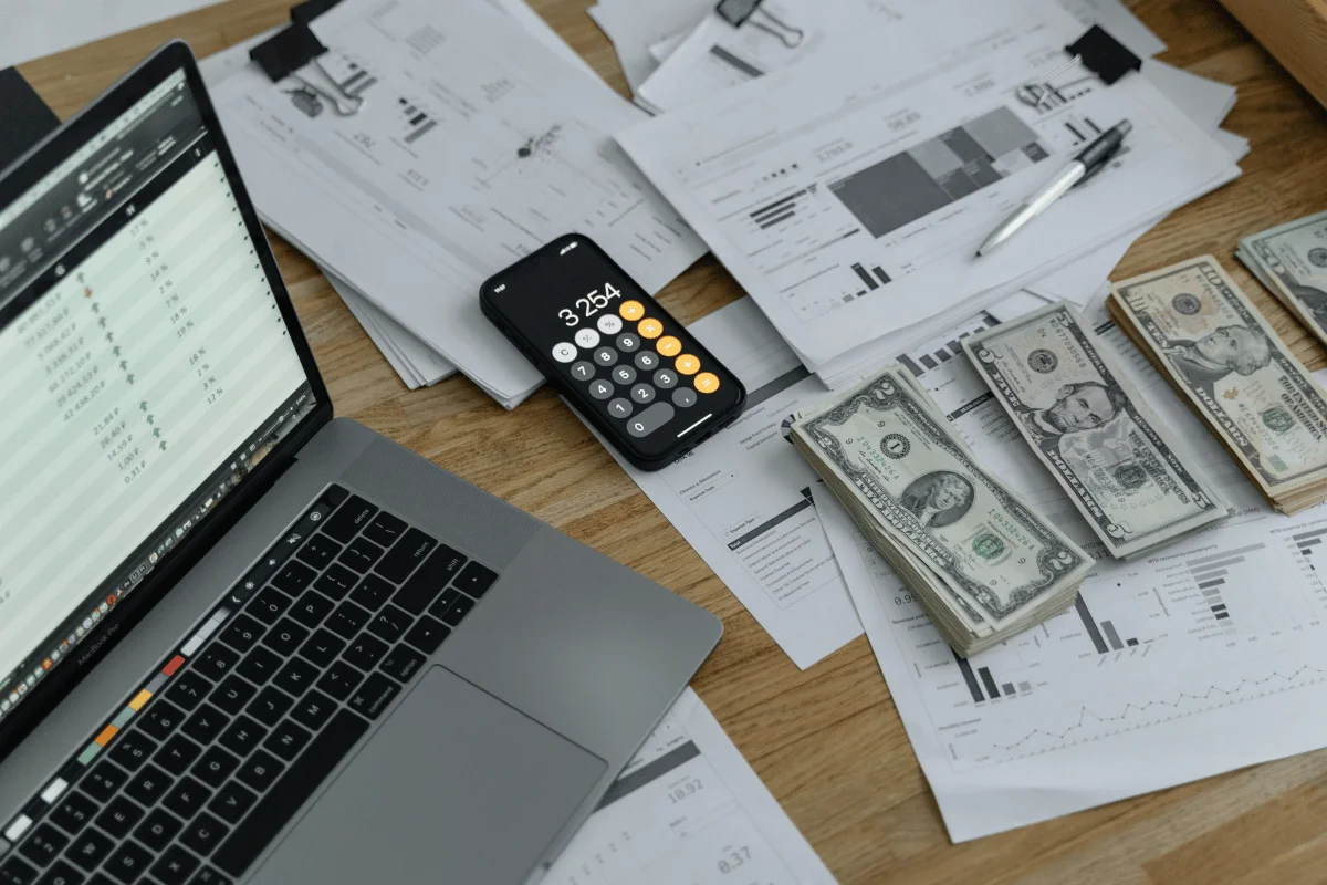 Financial audit desk setup featuring cash stacks, graphs, a laptop, and a smartphone calculator.