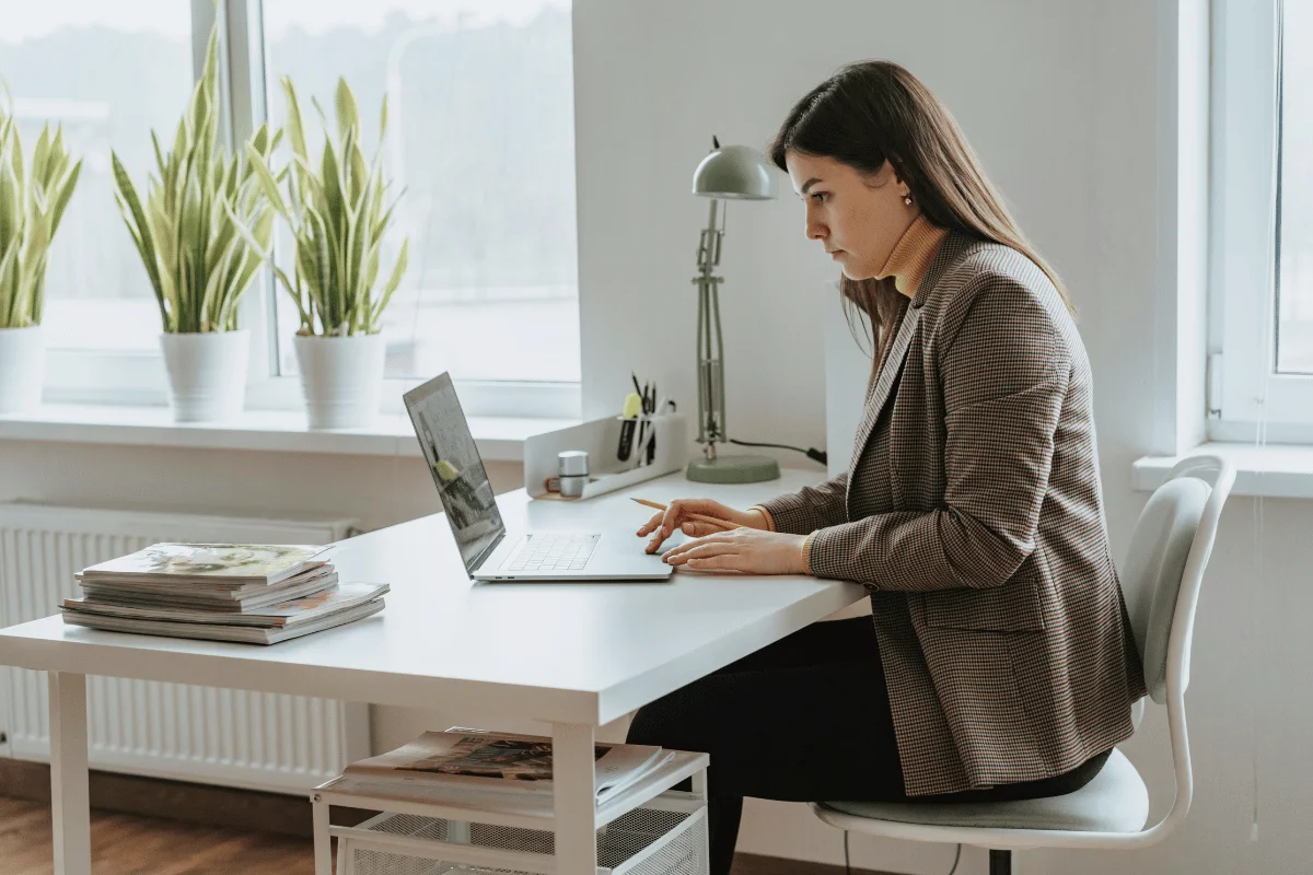 A person sits at a modern desk with a laptop, surrounded by potted plants and organized stationery, working in a bright office space.