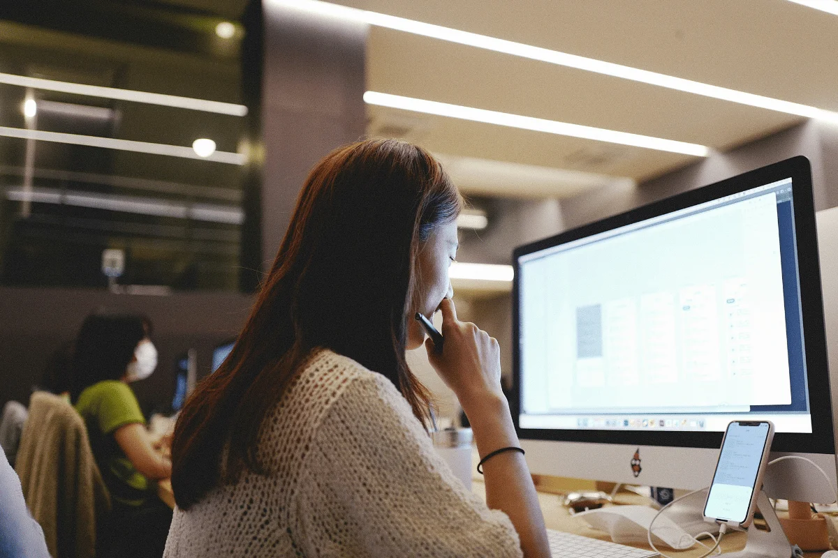 A woman in a knit sweater is focused on a computer screen in a modern office, holding a pen to her chin.