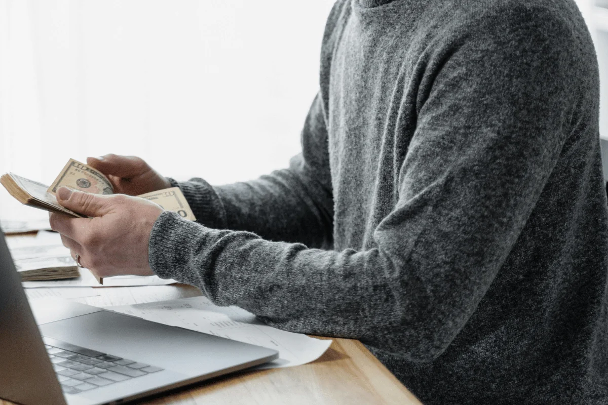A person in a gray sweater counts cash while sitting at a table with a laptop and papers.
