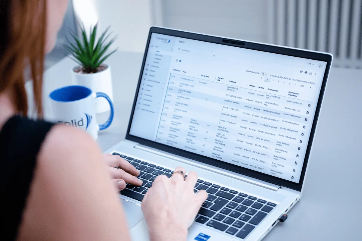 A person types on a laptop displaying a detailed spreadsheet, with a potted plant and a blue coffee mug in the background.