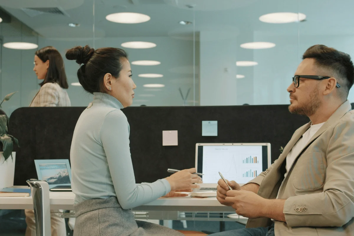A man and woman sit at a modern office table, engaged in discussion, with a laptop displaying charts.